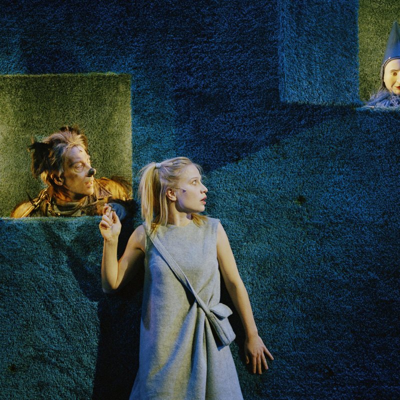 kvinna står mot blå vägg där två figurer sticker ut sina huvuden ur luckor 
