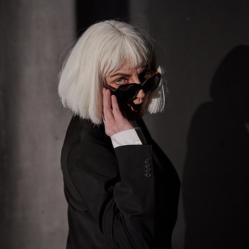 Kvinna i blond peruk tittar fram bakom svarta solglasögon
