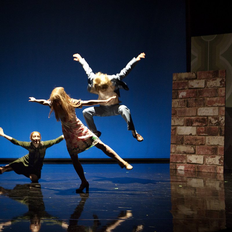 Tre personer hoppar och dansar på scen medan en man i underkläder står i utkanten