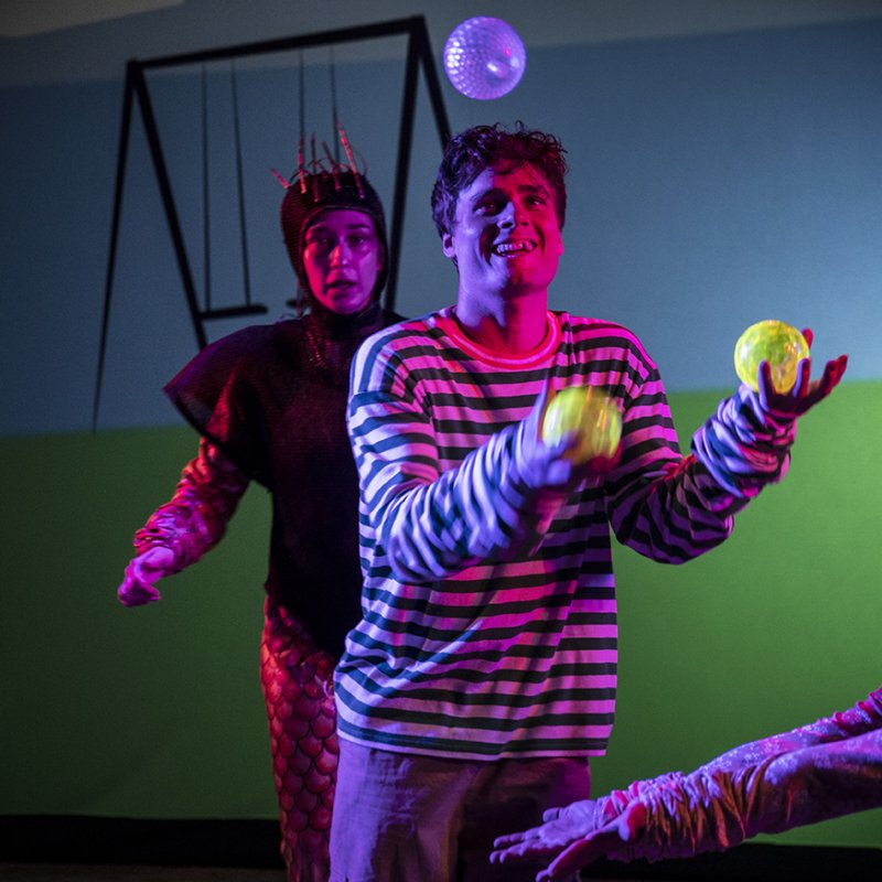 En kille med randig tröja jonglerar med tre bollar som lyser i mörkret medan en kvinna iklädd med en jumpsuit och en svart luva och väst står bakom honom och tittar på honom och en annan kvinna står till höger av killen och försöker fånga in bollarna.  
