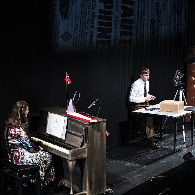 En smal teaterscen med en pianist och en skådespelare som sitter framför ett bord med olika föremål och bilder. I bakgrunden står en stor koffert.