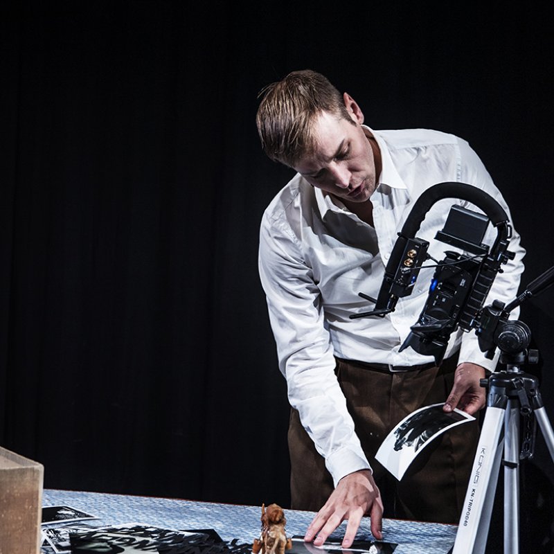 Skådespelaren visar olika fotografier och föremål via en filmkamera på ett staffli som är riktad mot bordet med föremålen.