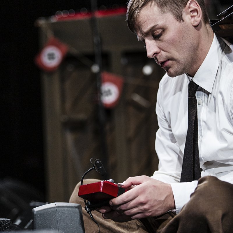Skådespelaren sitter i skräddarställning på scengolvet och spelar på ett tumpiano. 