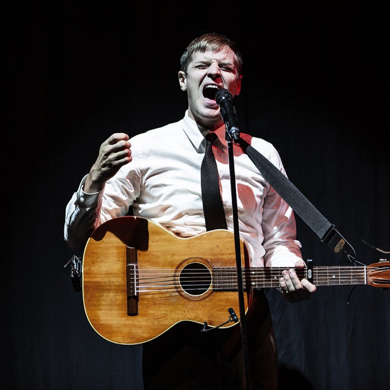 Skådespelaren spelar gitarr och sjunger med anspänt ansiktsuttryck.