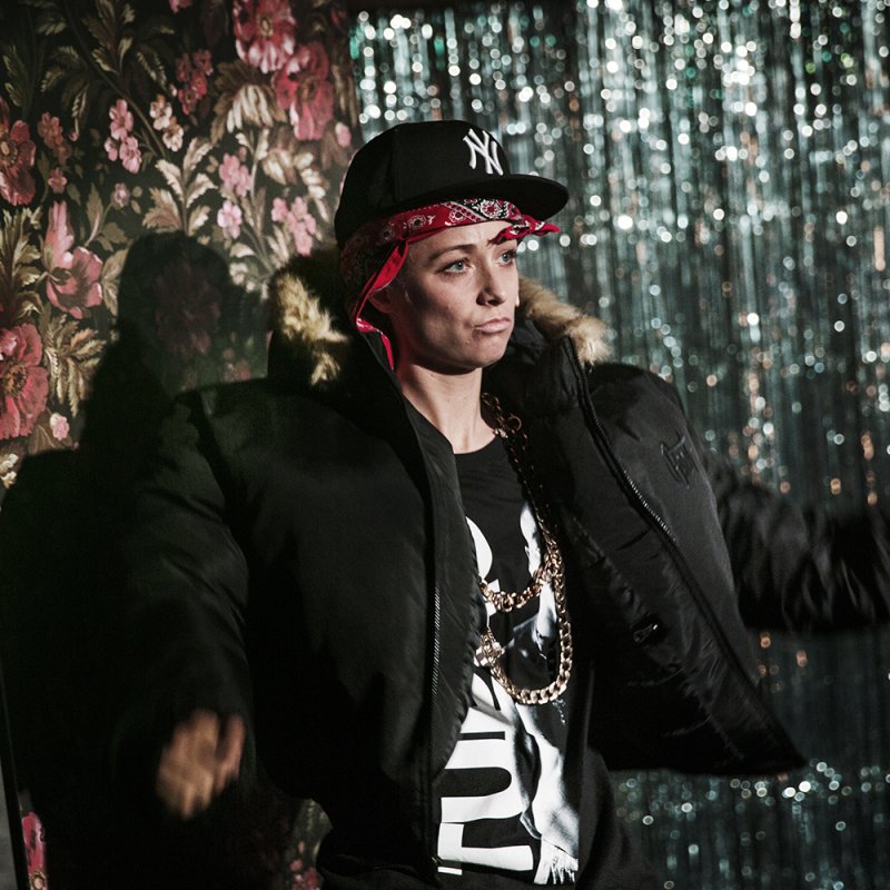 Tjej i svart keps och svart jacka i hip hop-stil dansar fram på scenen framför en glitterridå.