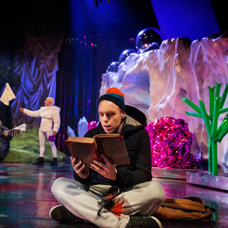 En pojke sitter och läser ur en bok och i bakgrunden visas ett fantasilandskap med udda figurer.