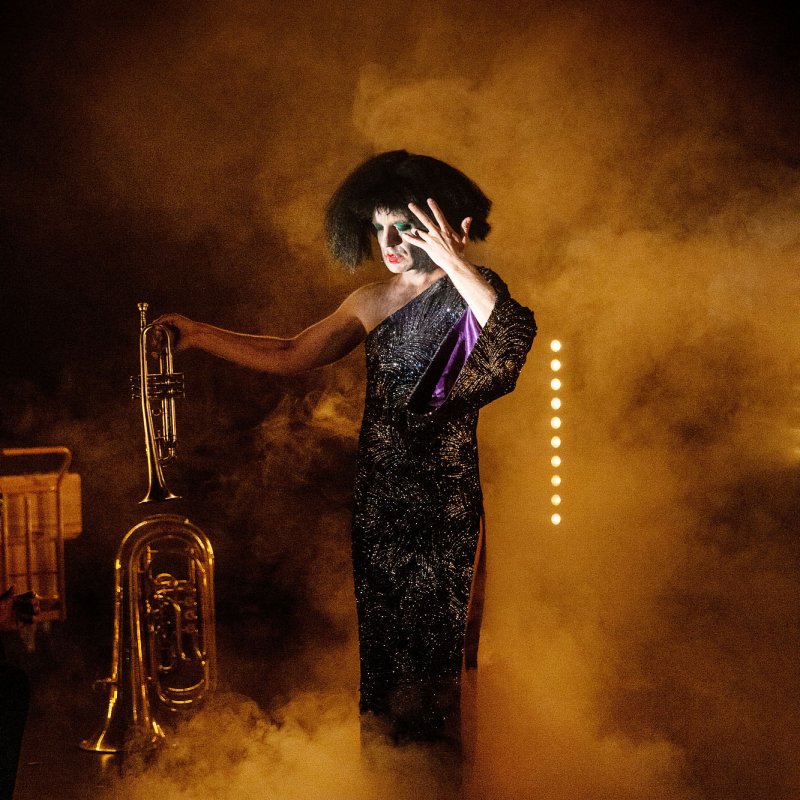 Person i glitterklänning och svart peruk i teaterrök, håller i en trumpet