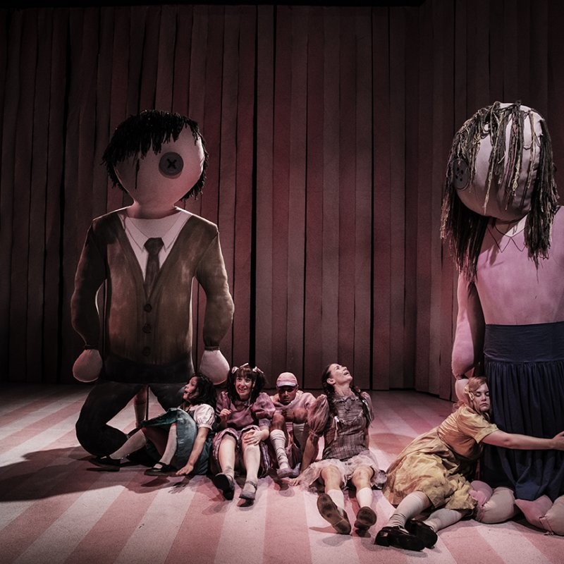 Fem barn sitter på golvet och tittar upp mot 2 stora dockor, en som föreställer en man och en som föreställer en kvinna. Ett av barnen kramar den kvinnliga dockan, 