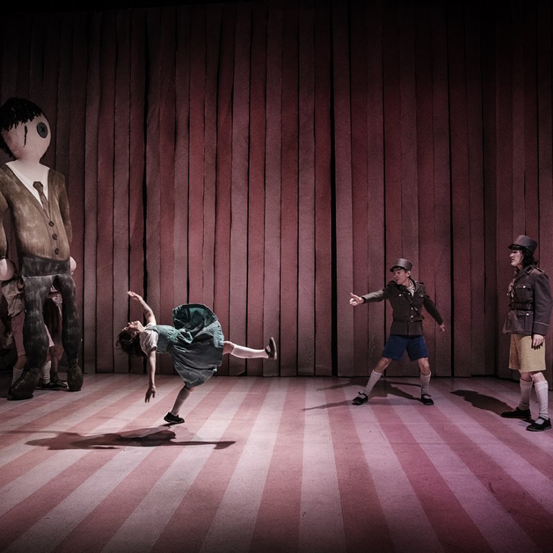 En flicka dansar i mitten av en scen som en rosa och randig medan en pojke och en soldat tittar på henne. Åt vänster av dem står även en stor docka som förställer en man. 
