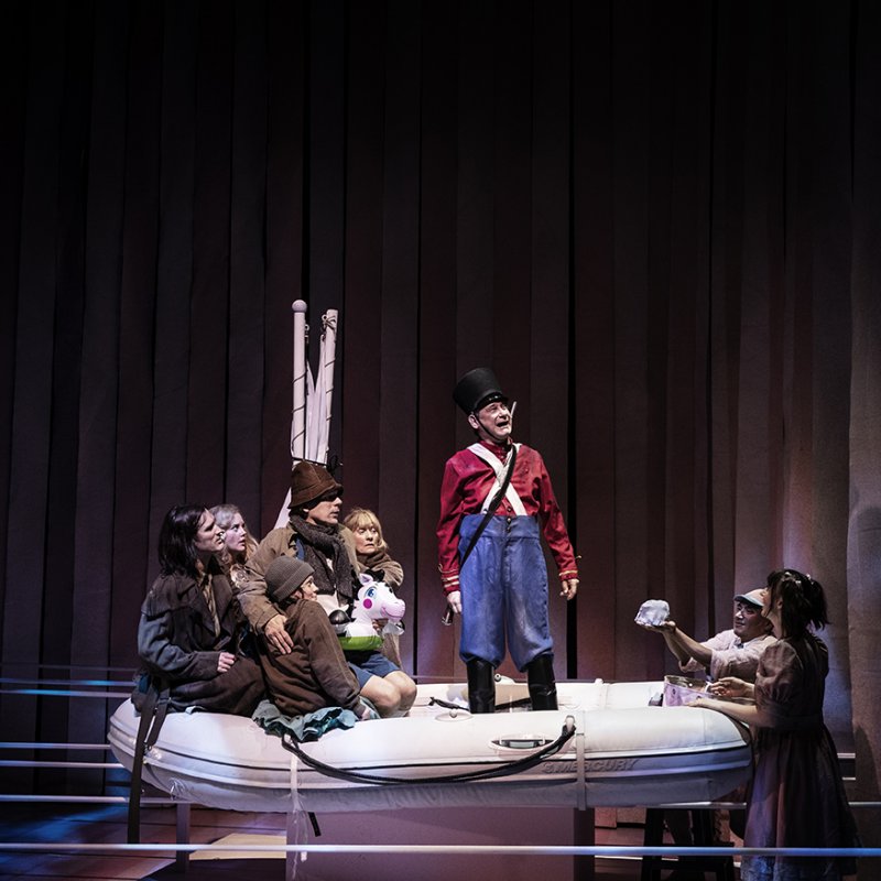 Fyra barn iklädda i bruna och mörka kläder sitter på en båt där det står en man klädd med röda och blåa kläder som föreställer tennsoldaten, Det står även ett barn vid högersida av båten. 