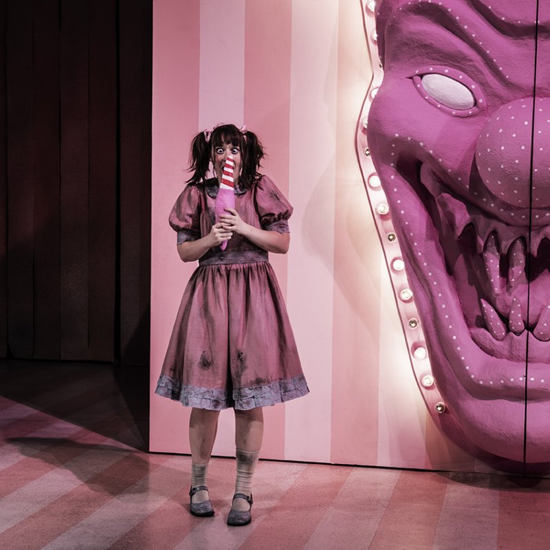 En flicka i rosa kläder står åt vänster av scenen och slickar på en falsk polkagris och stirrar på ett hus med ett monsteransikte utanför. 