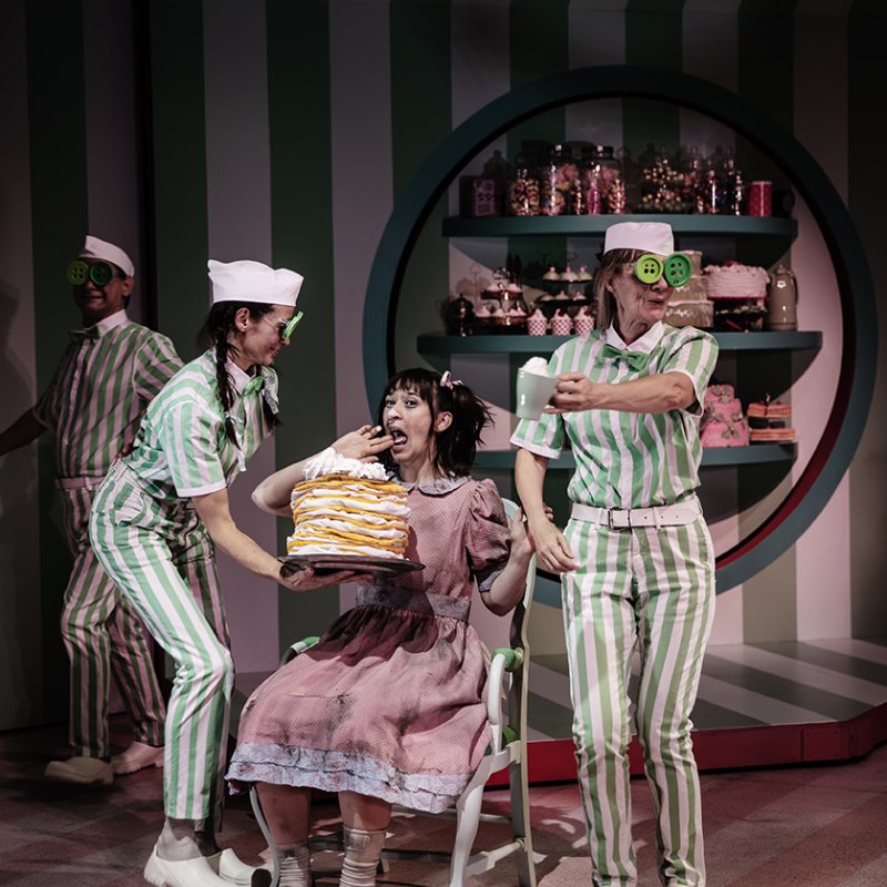två personer med grön och vita dräkter och vita mössor står framför en väg med många tårtor och godis och ger en tårta till en flicka som sitter i en stol. 