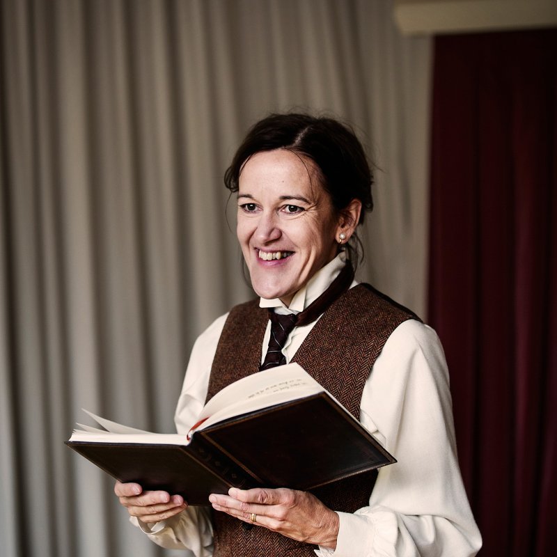 Kvinna i strikt kostym av äldre snitt håller en upplagen bok och skrattar med blicken i fjärran.