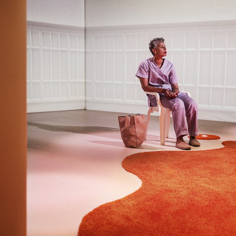 Kvinna sitter i vit stol till höger. Orange matta på golvet.