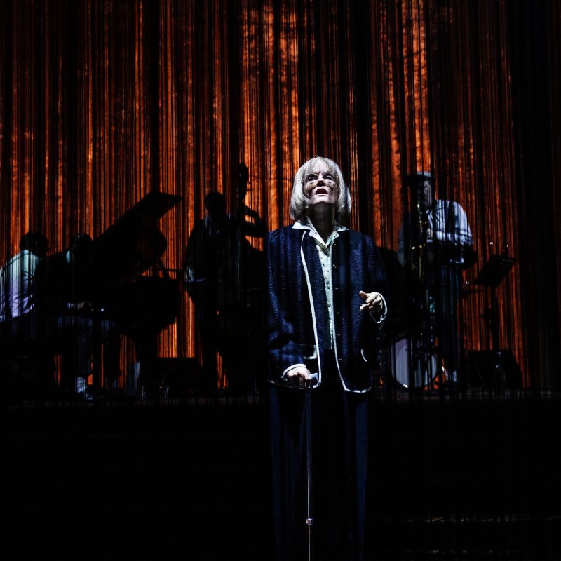 En äldre Monica står upp framför ett draperi bakom vilket ett band står och spelar musik.