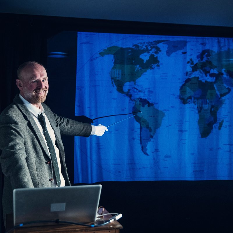 En man kostym föreläser framför en världskarta.