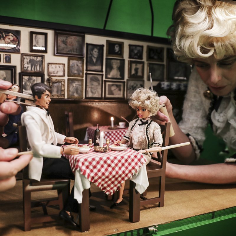 Två dockor äter middag tillsammans i en dockskåpsrestaurang. En skådespelare håller i en av dockorna.