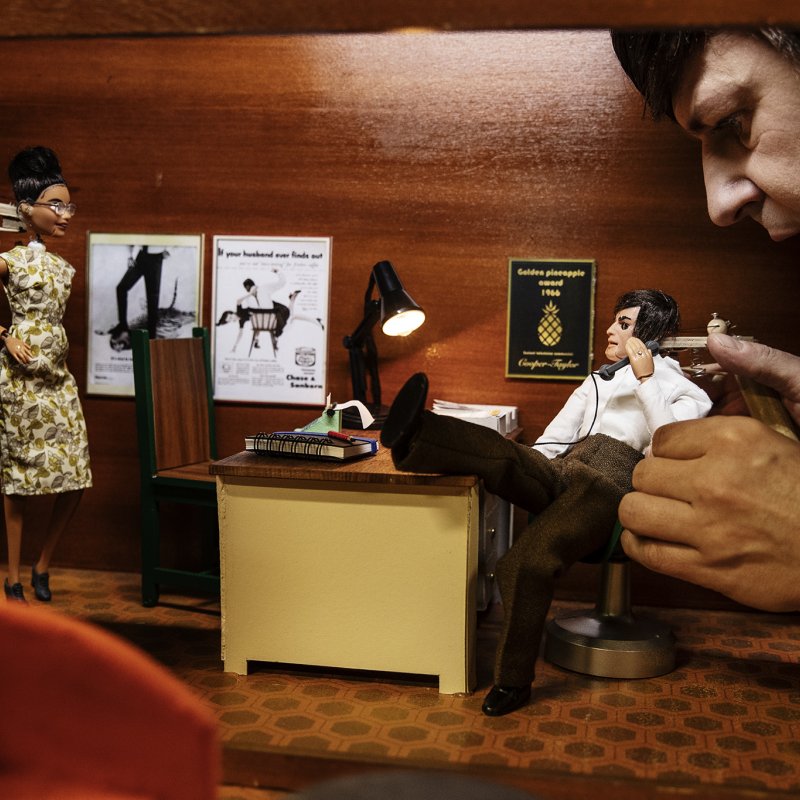 Två dockor pratar med varandra i kontorsmiljö. En skådespelare håller i en av dockorna.