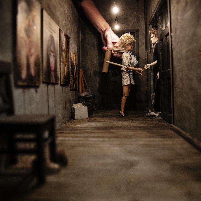Två dockor står i dörröppningen till en lägenhet, i en lång korridor