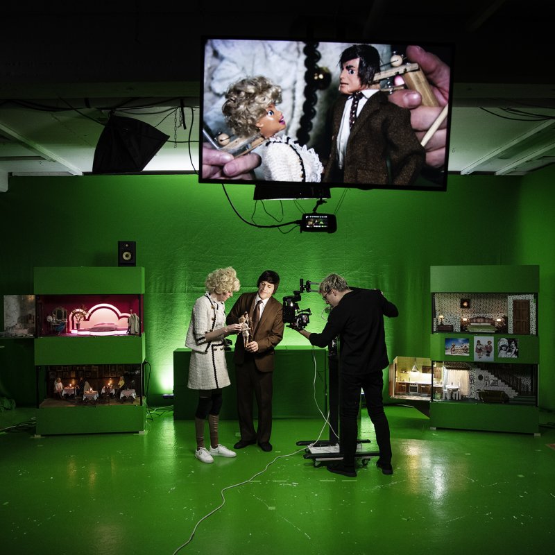 Ett stort grönt rum fullt med dockskåp. Två skådespelare och en filmare står mitt i rummet.