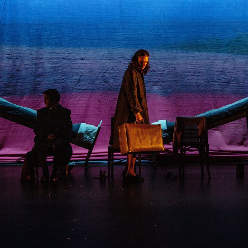 Kvinna i mitten av scenen med resväska i handen. Två sängar lyfts på var sida om henne. Rosablått ljus.