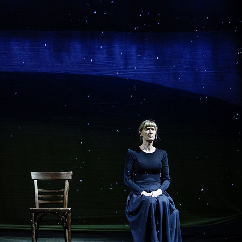 En kvinna i svart klänning sitter på en stol. En tom stol till vänster om henne. Blått scenljus och stjärnhimmel.