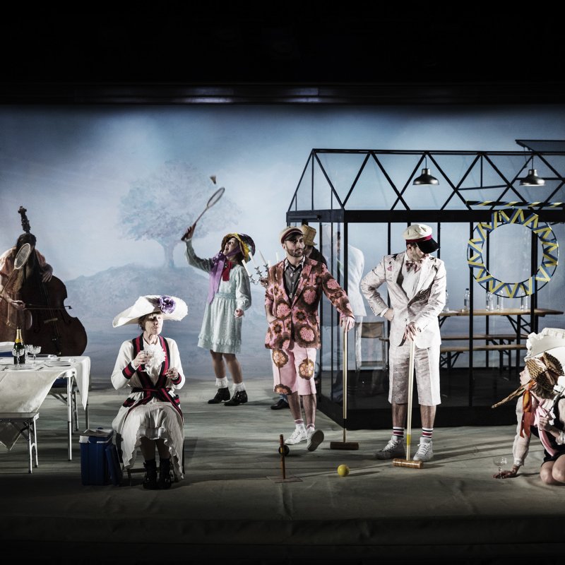 Skådespelare i märkliga kläder sysselsatta med picknick-aktiviteter. I bakgrunden står ett växthus på scenen.