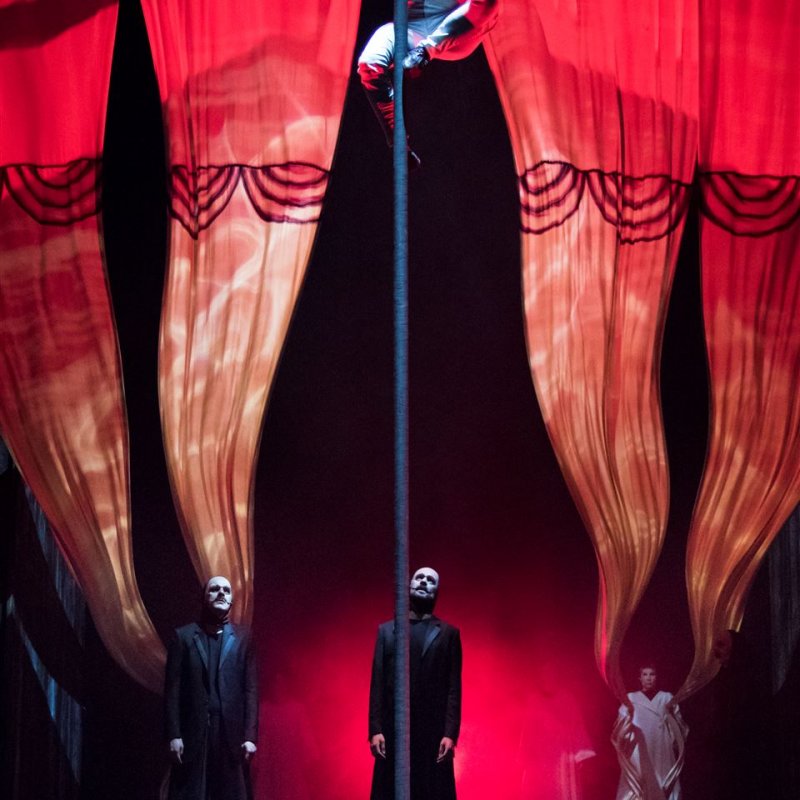 En kvinna håller sig fast högst uppe på en stång. Två mörkklädda figurer står under och ser upp på henne och en vitklädd person håller i ett par röda draperier som böljar