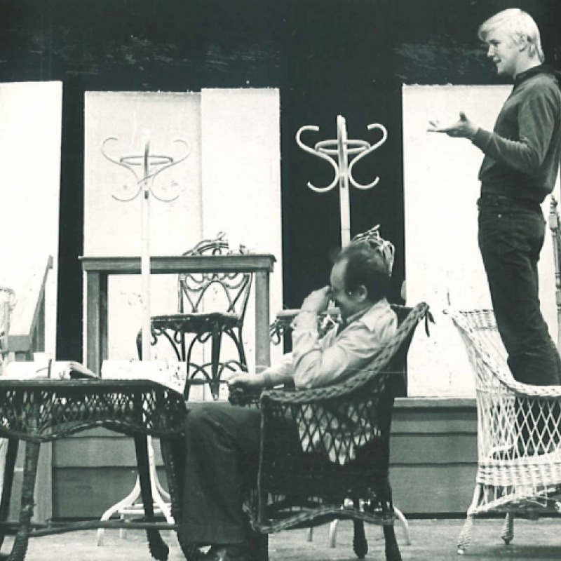 En ung man står på en korgstol och gestikulerar medan personer runt omkring skrattar