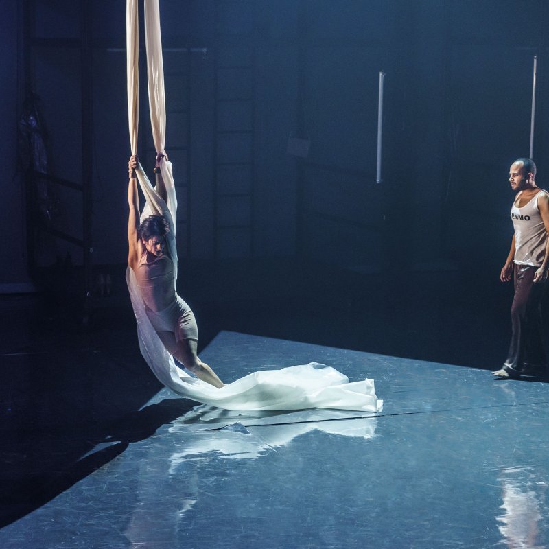 Kvinna utför cirkuskonst i ett tygdrapperi medan en man står vid sidan av och betraktar en annan person som har ryggen mot.