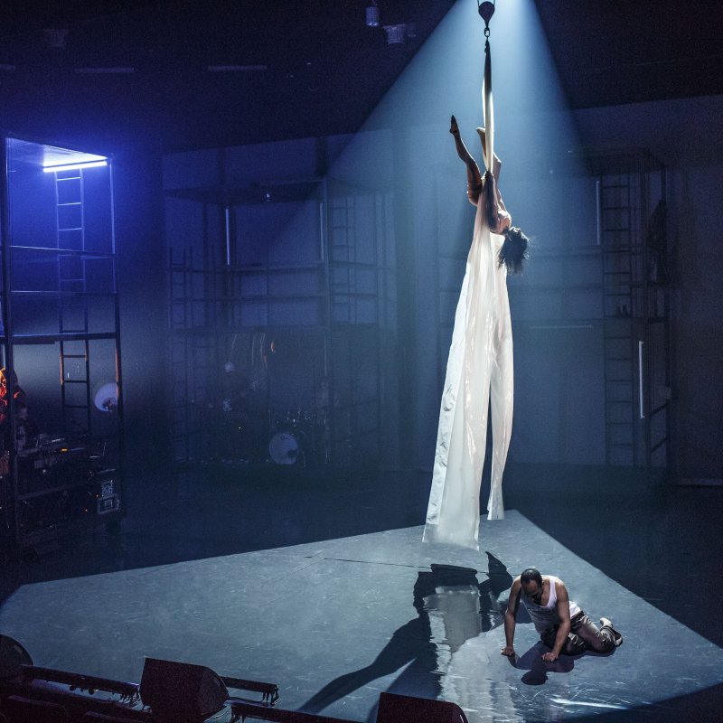 En cirkusartist hänger i ett långt plastskynke från taket medan en man sitter på knä på scengolvet under.