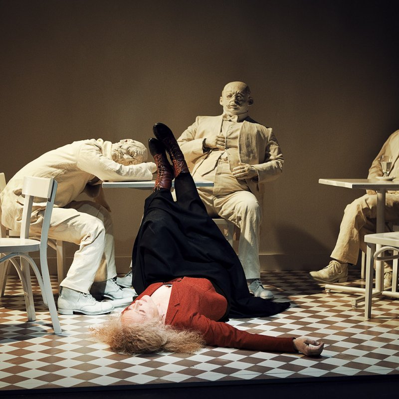 Kvinna ligger på golvet medan tre vitklädda figurer sitter kring ett bord