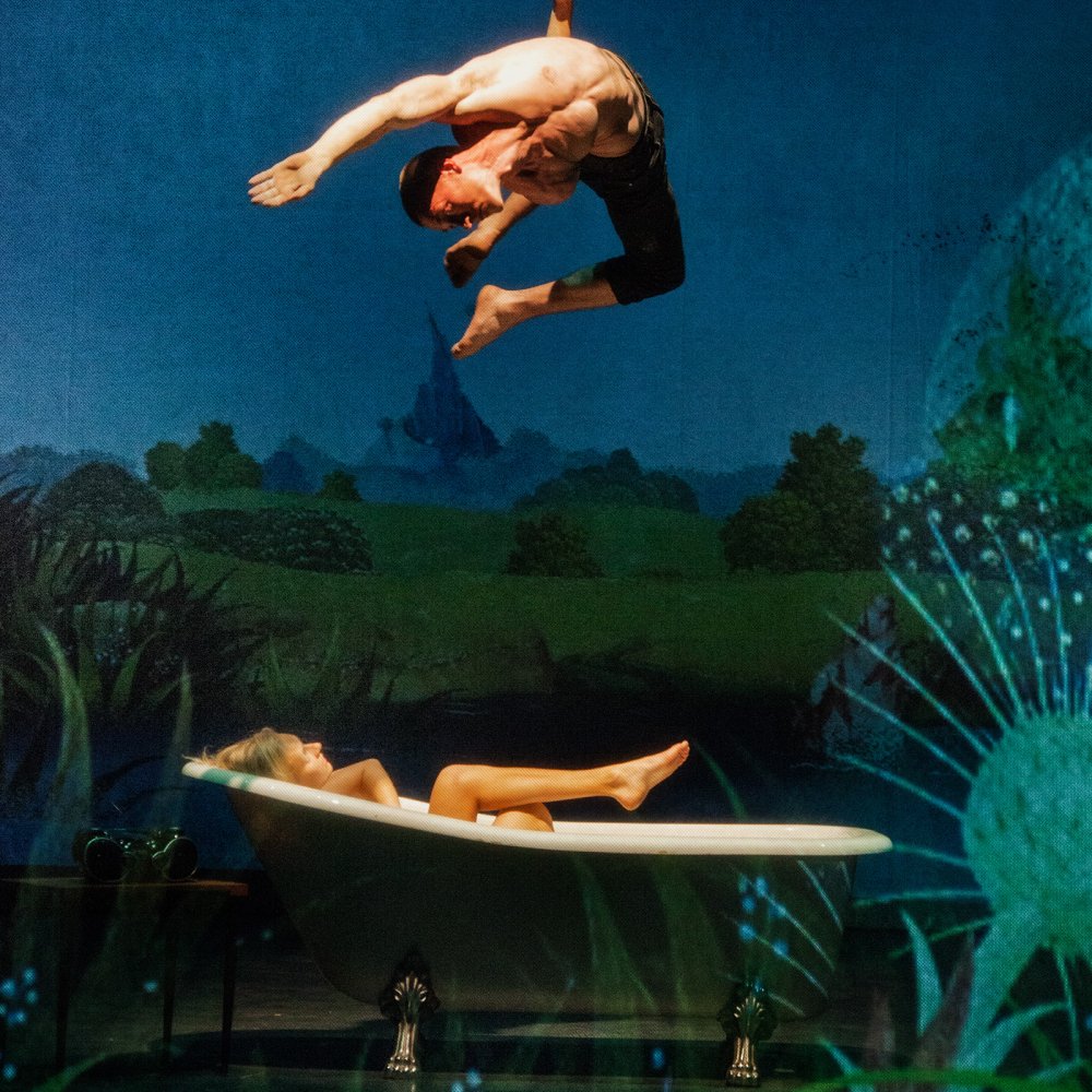 En trapetskonstnär hänger över en kvinna som ligger i ett badkar