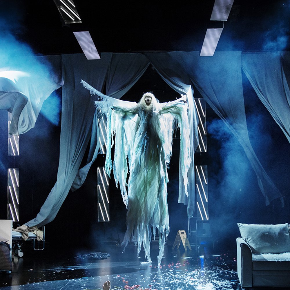En ängel flyger in över scenen omgiven av blå draperier. 