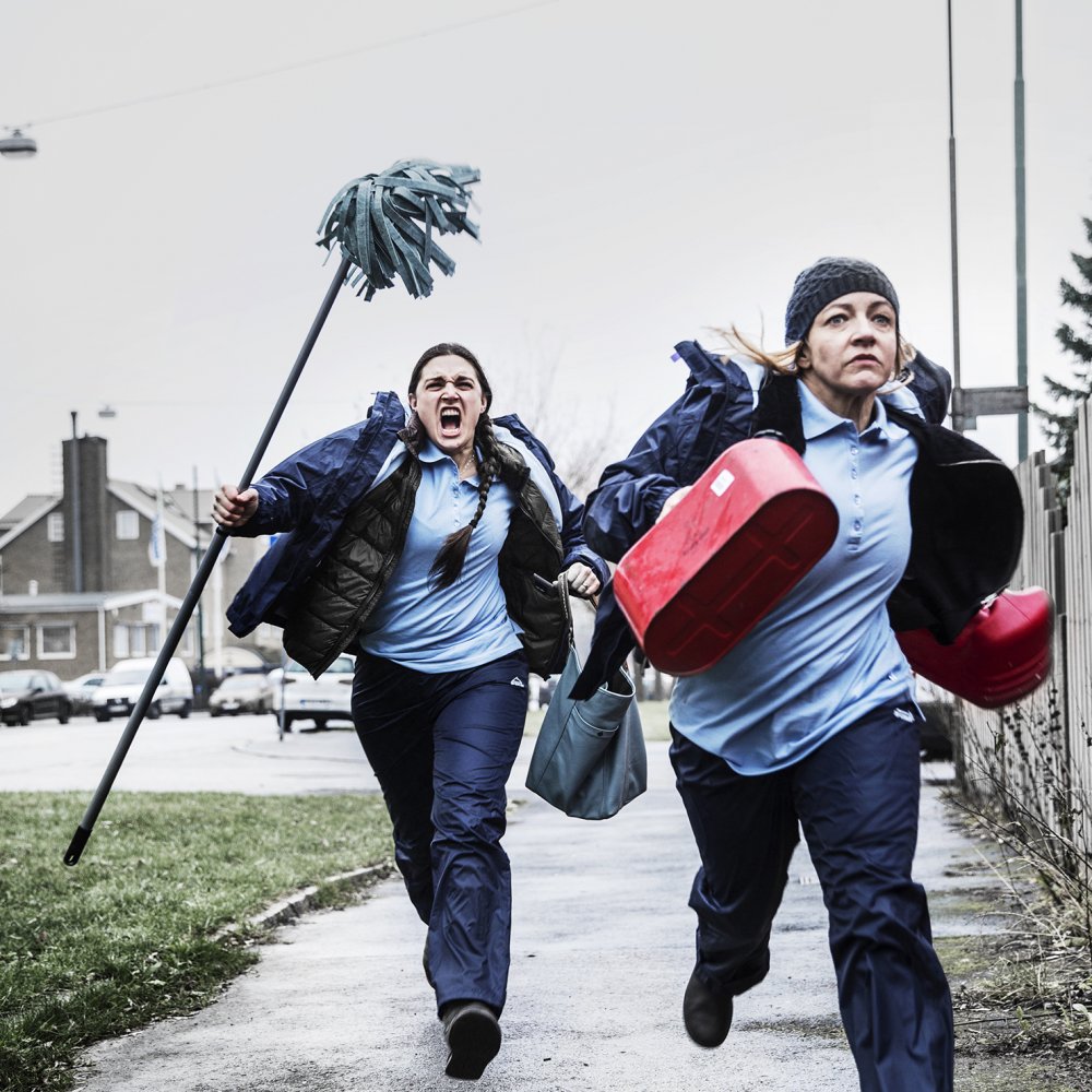 Två kvinnor i blå socialtjänstkläder springer i panik med städmopp och bensindunkar i händerna