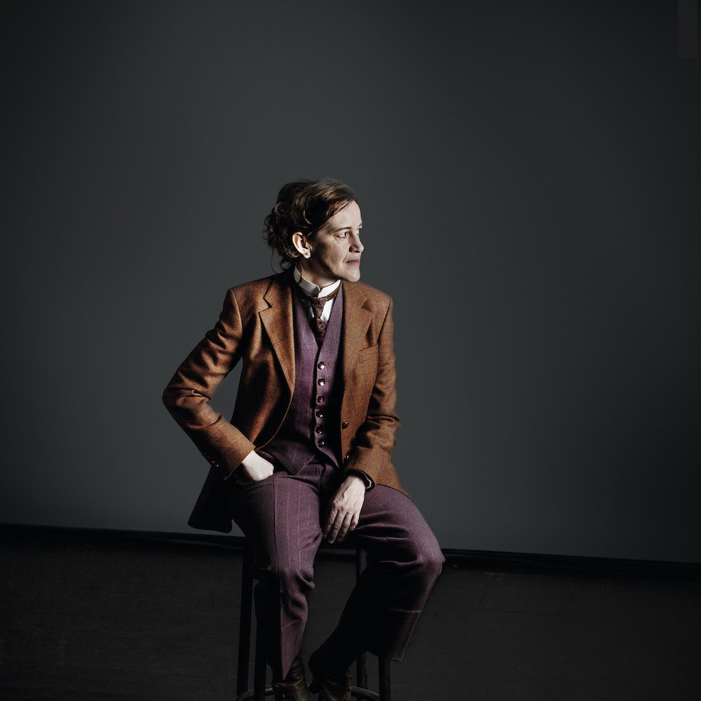 Skådespelaren Cecilia Lindqvist sitter på en pall i en full kostym och tittar fundersamt åt sidan.