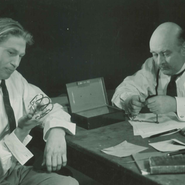 Två män sitter bland en mängd dokument och andra objekt vid bord